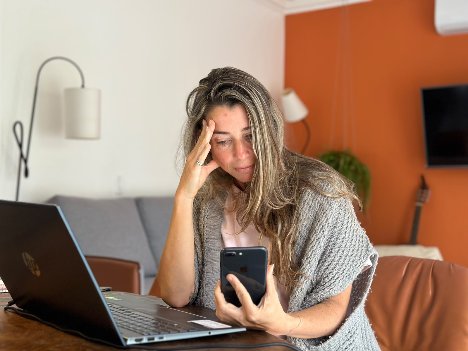 Ana Tahan sentada à mesa, com a mão na testa, olhando o celular ao lado do notebook